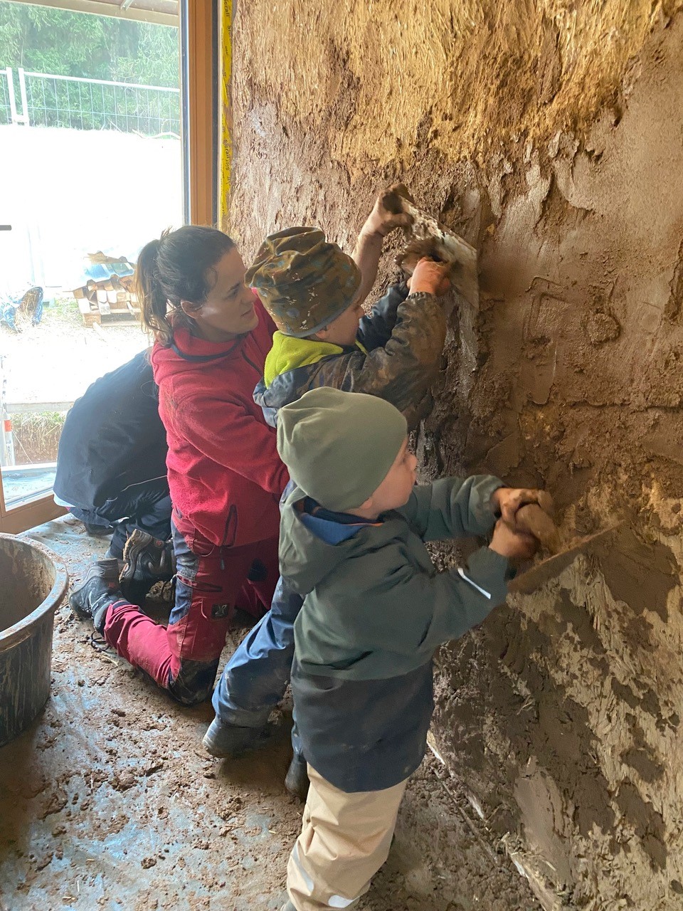 Im Waldkindergarten Pinzberg sind eine Frau und zwei kleine Waldwichtel damit beschäftigt, Lehm auf eine Innenwand aufzutragen. Sie nehmen an einer praktischen Verputzaktion teil, die ihr nachhaltiges Modulsystem und ihre Liebe zum Lernen durch Handeln widerspiegelt. at Hochschule Coburg