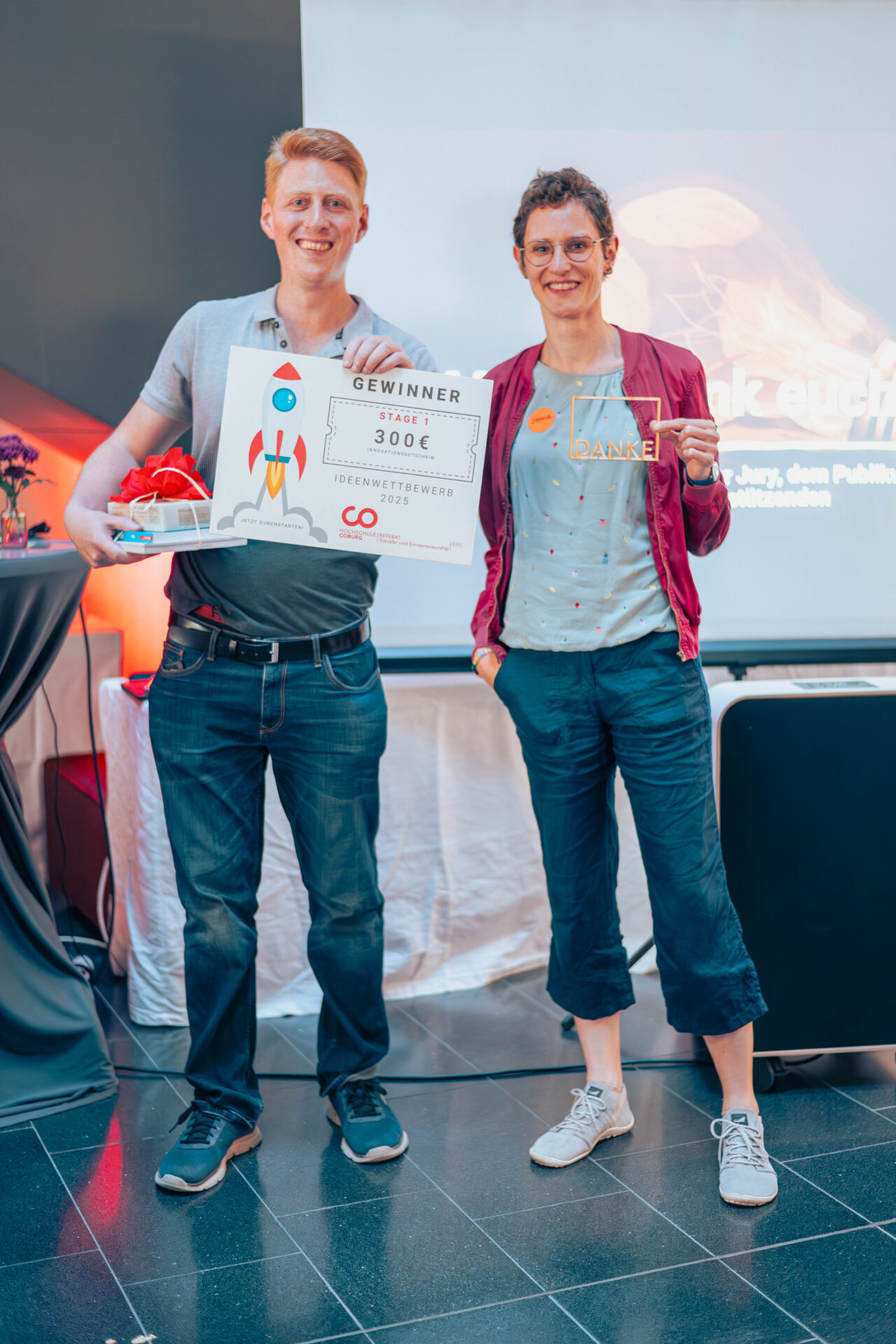 Zwei Menschen stehen lächelnd in einem Raum. Beim Finale des Ideenwettbewerbs an der Hochschule Coburg hält der eine ein "Gewinner"-Schild mit "300€" und einer Raketen-Illustration in der Hand, dazu einen Preiskorb. Der andere trägt ein "DANKE"-Shirt. Hinter ihnen stehen eine Leinwand und ein Tisch. at Hochschule Coburg