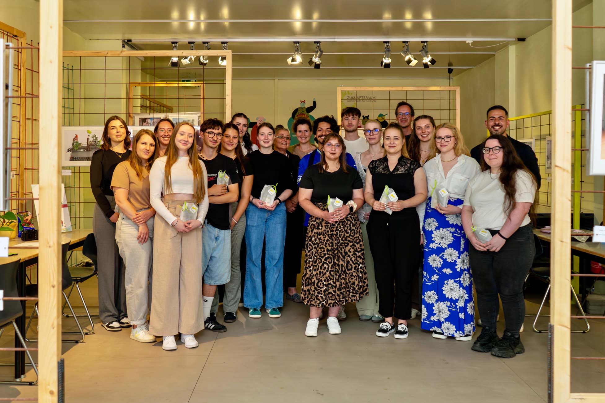 Eine Gruppe von etwa zwanzig Coburger Studierenden steht in einem Innenraum zusammen und lächelt in die Kamera. Einige halten kleine Geschenktüten in der Hand. Im Hintergrund sind Kunstwerke und Oberlichter in einem modernen, kreativen Raum zu sehen, die ihre Rolle bei der Gestaltung von Stadt und Zukunft widerspiegeln. at Hochschule Coburg