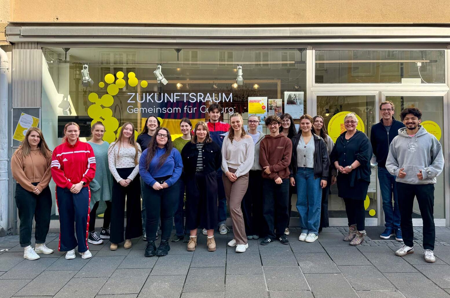 Eine Gruppe von sechzehn Personen steht lächelnd vor einer Schaufensterfront mit großen Fenstern und gelber Seifenblasendekoration. Auf dem Schild über dem Schaufenster steht "Zukunftsraum - Gemeinsam für Coburg". Studierende in Coburg engagieren sich, um die Zukunft zu gestalten. at Hochschule Coburg