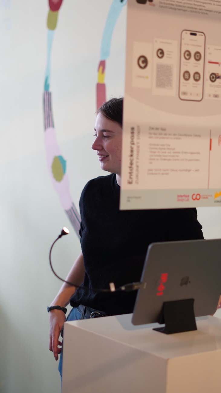 Eine Person in einem schwarzen Hemd steht neben einem Podium mit einem Laptop, teilweise verdeckt durch ein großes Plakat darüber. Im Hintergrund ist eine helle Wand mit einem farbenfrohen, abstrakten Wandgemälde zu sehen - eine inspirierende Szene über die Studierenden Coburgs, die die Zukunft gestalten. at Hochschule Coburg