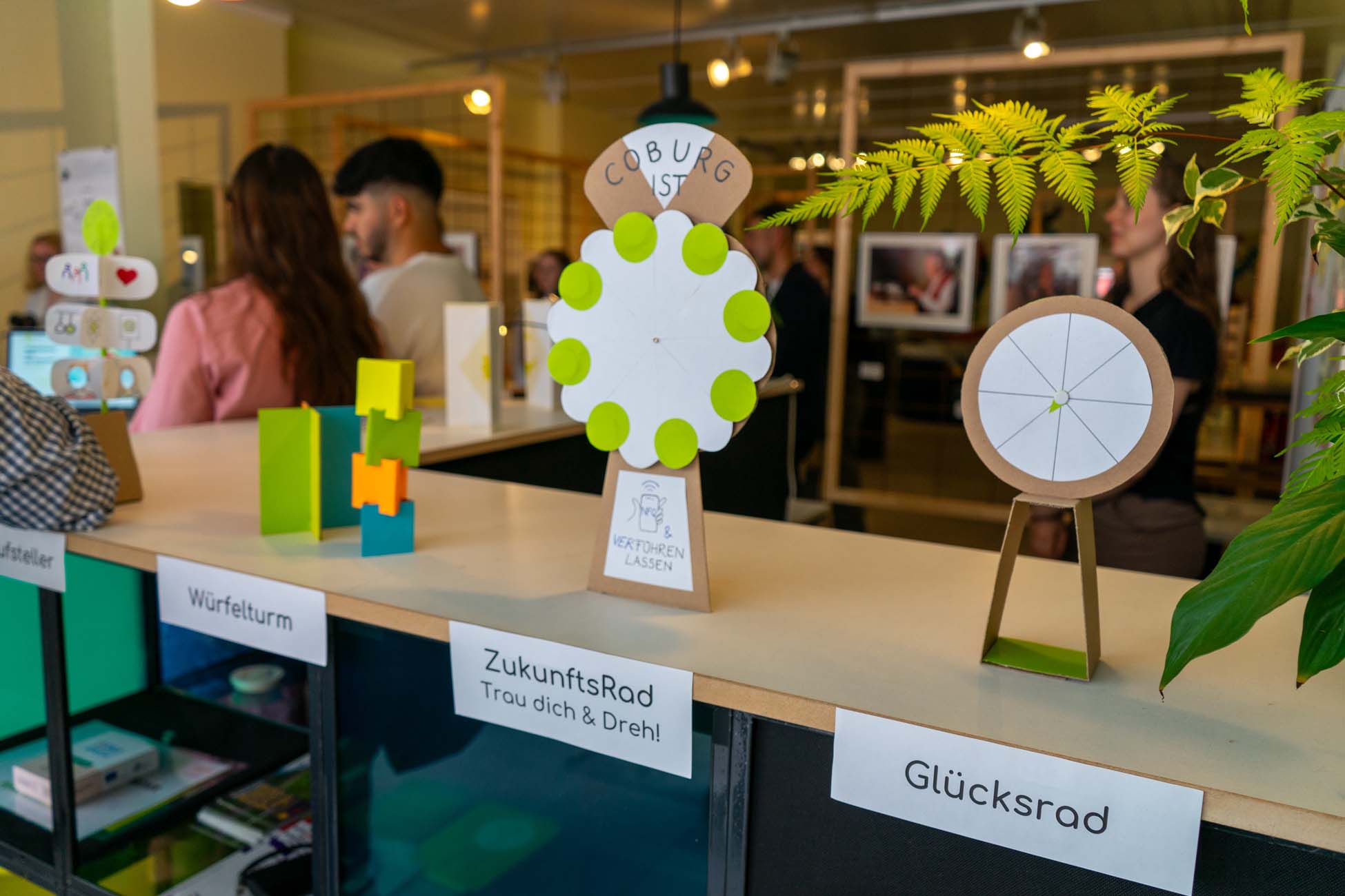 Ein Schautisch in Coburg zeigt interaktive Spiele für Studierende, darunter bunte Klötzchentürme und zwei sich drehende Räder mit der Aufschrift "ZukunftsRad" und "Glücksrad", während im Hintergrund über Stadtgestaltung diskutiert wird. at Hochschule Coburg