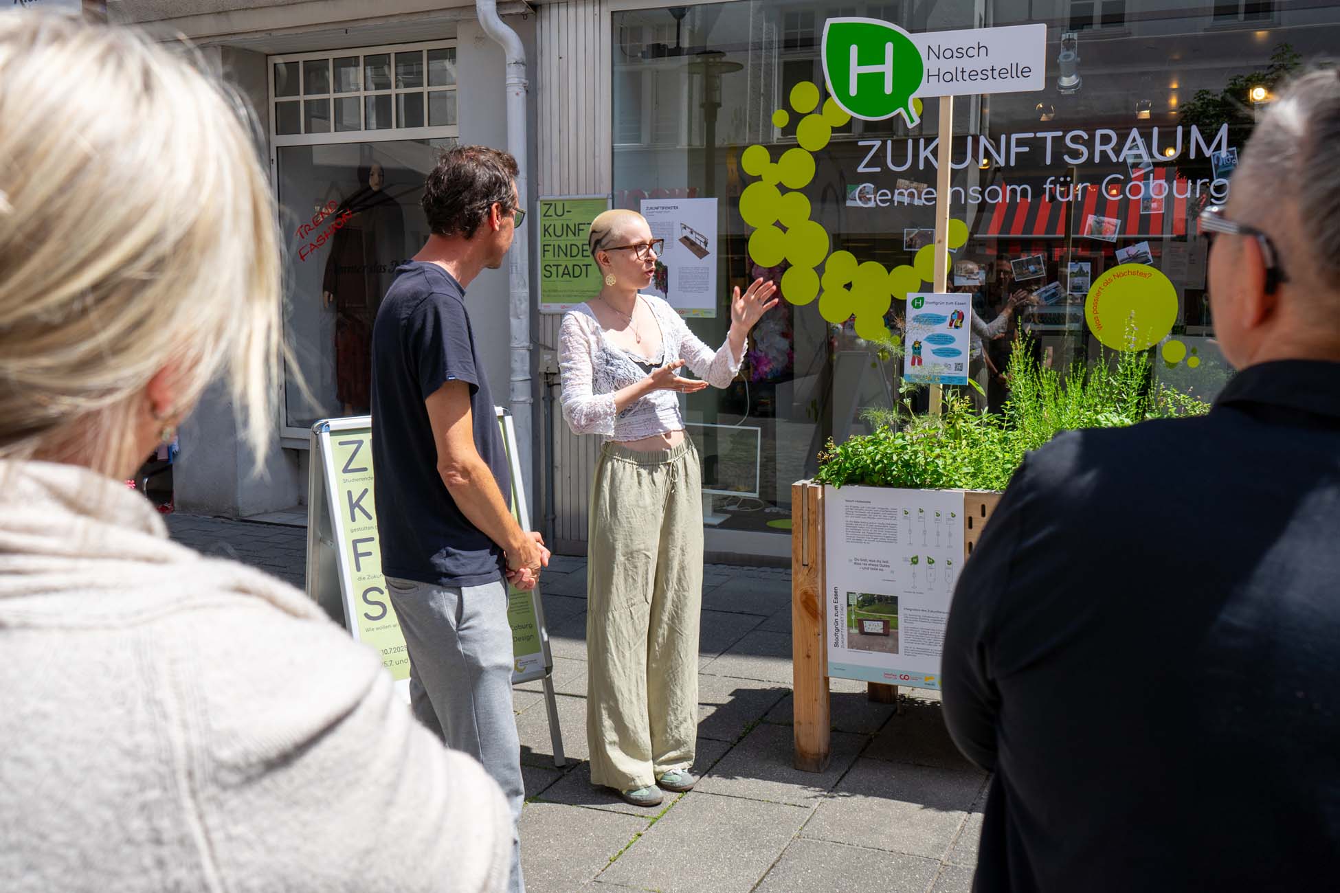 Eine Frau spricht und gestikuliert zu einer kleinen Gruppe vor einem Schaufenster mit der Aufschrift "ZUKUNFTSRAUM - Gemeinsam für Coburg". Die Studierenden hören zu, während in einem Pflanzgefäß neben ihr frische Kräuter in der Sonne auf der Straße wachsen. at Hochschule Coburg