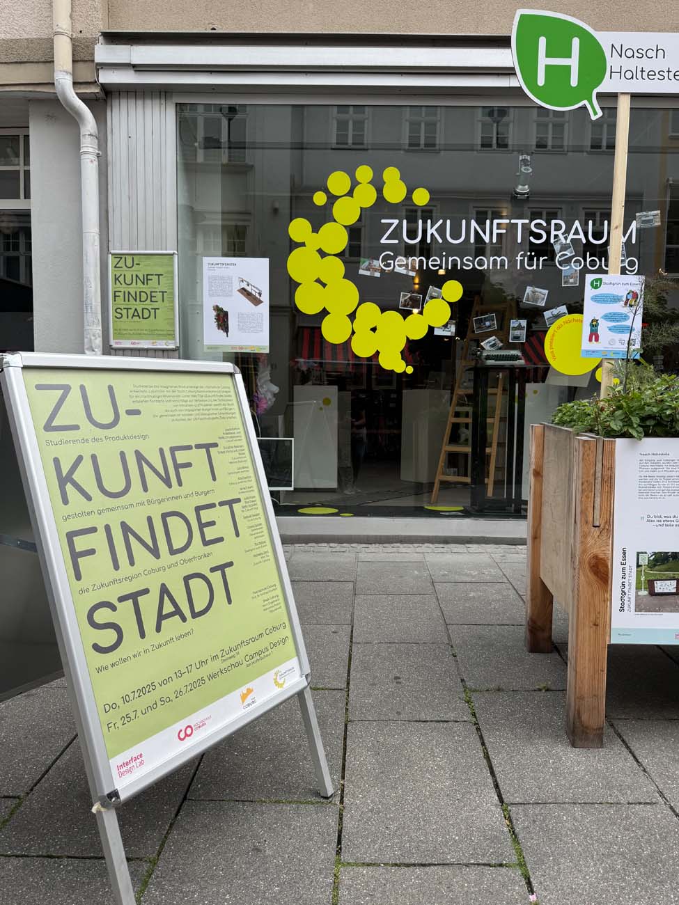 Ein Schaufenster mit dem Schriftzug "Zukunftsraum - Gemeinsam für Coburg" und großen gelben Punkten auf dem Schaufenster. Auf einem Plakat steht "Zukunft findet Stadt". Der Raum lädt Studierende Coburg zu städtischen Veranstaltungen ein, darüber ein grünes "Nasch Haltestelle"-Schild. at Hochschule Coburg