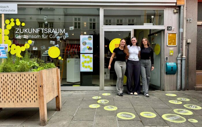 Drei Frauen stehen lächelnd vor dem modernen Schaufenster "Zukunftsraum - Gemeinsam für Coburg", das die Stadtentwicklung präsentiert. Kreisförmige gelbe Aufkleber zieren das Schaufenster und den Gehweg, links ist ein großer Pflanzkübel zu sehen. at Hochschule Coburg