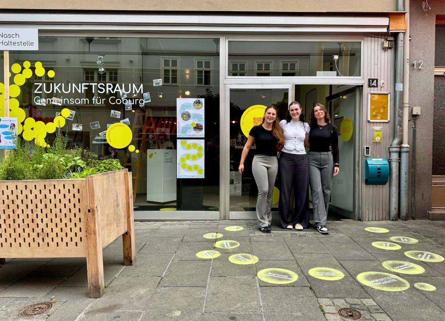 Drei Frauen stehen lächelnd vor dem modernen Schaufenster "Zukunftsraum - Gemeinsam für Coburg", das die Stadtentwicklung präsentiert. Kreisförmige gelbe Aufkleber zieren das Schaufenster und den Gehweg, links ist ein großer Pflanzkübel zu sehen. at Hochschule Coburg