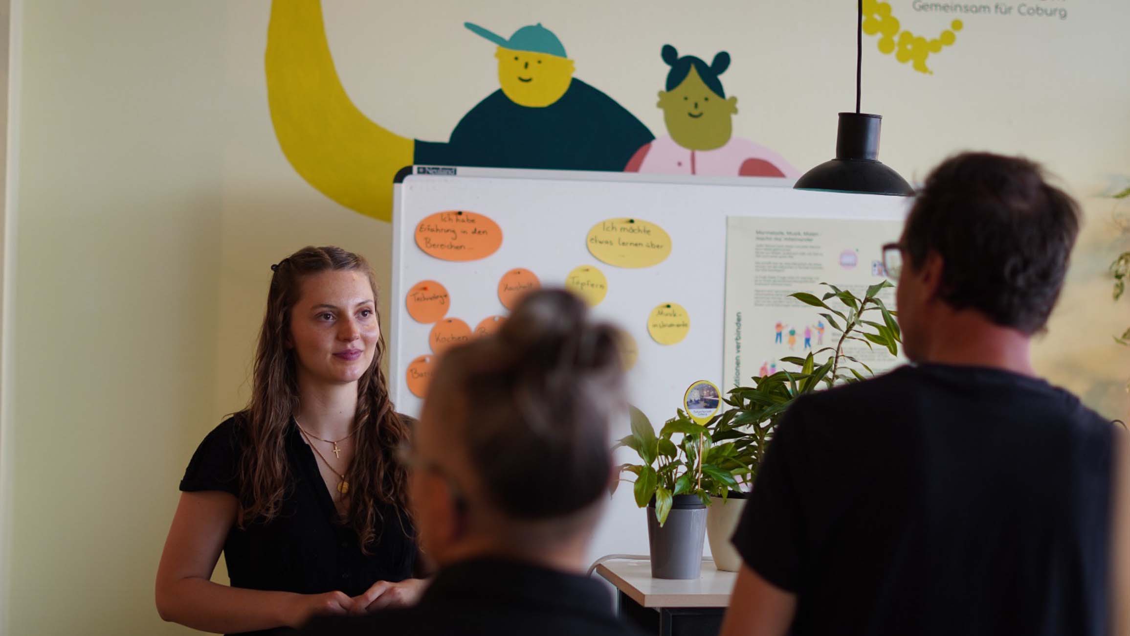 Eine Frau steht neben einem Whiteboard mit bunten Haftnotizen und spricht mit zwei Studierenden. Im Hintergrund ist ein Wandgemälde mit Cartoon-Figuren zu sehen, das den kreativen Geist von Stadt gestalten in Coburg widerspiegelt, und auf dem Tisch daneben steht eine Topfpflanze. at Hochschule Coburg