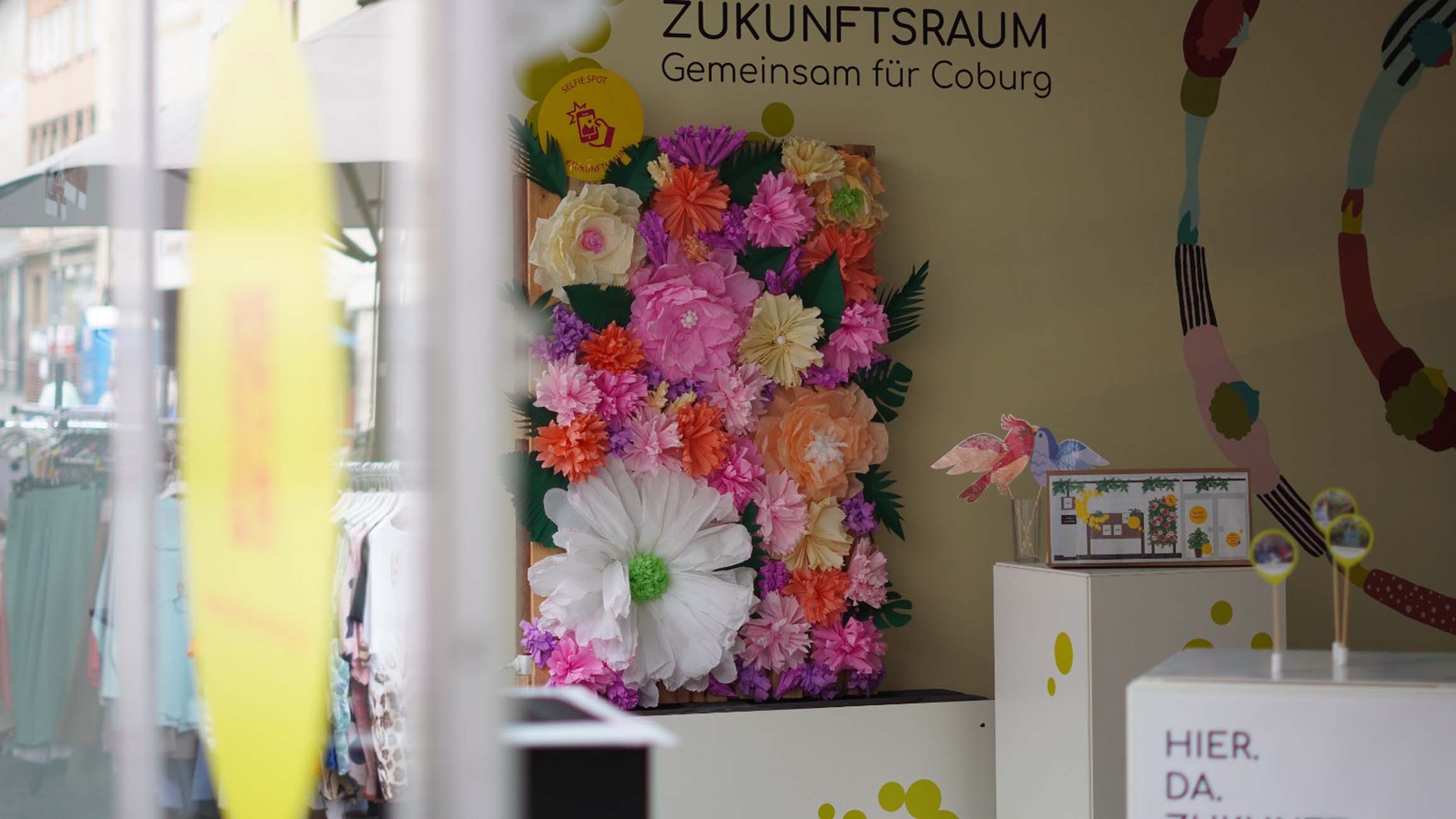 Eine Schaufensterauslage mit einem bunten Arrangement aus großen Papierblumen, einem kleinen Papiervogel und einem Schild mit der Aufschrift "ZUKUNFTSRAUM Gemeinsam für Coburg - Stadt gestalten mit Studierende Coburg" im Hintergrund. Draußen sind einige Kleiderständer zu sehen. at Hochschule Coburg