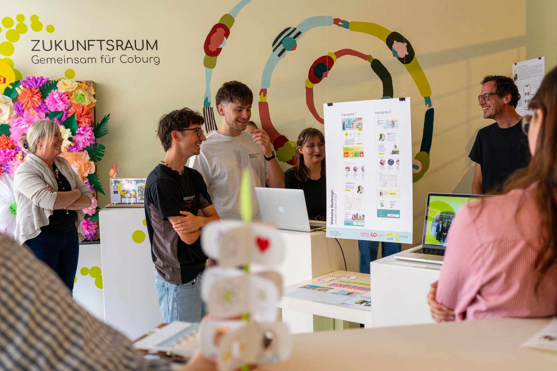 Eine Gruppe von Menschen, darunter auch Studierende, steht lächelnd um eine Schautafel mit Grafiken in einem hellen, mit Blumen und bunter Wandkunst geschmückten Raum. Auf dem Schild steht "Zukunftsraum - Gemeinsam für Coburg" und es werden Ideen für die Stadtgestaltung vorgestellt. at Hochschule Coburg