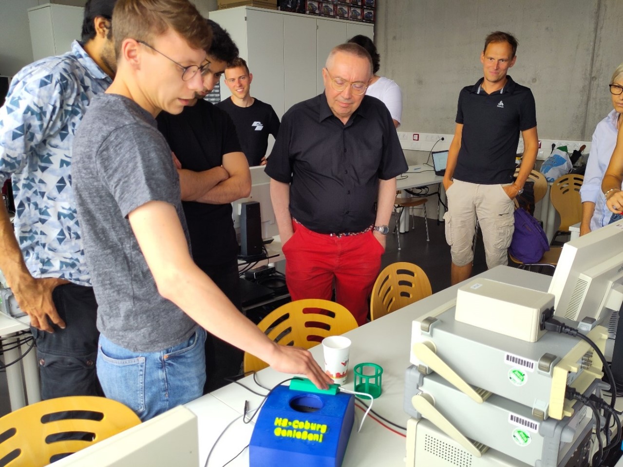 Eine Gruppe von Menschen versammelt sich um einen Tisch und beobachtet eine Person, die ein Gerät mit der Aufschrift "Hi-Coburg Onion01" neben alten Computern bedient. Eine Person hält einen Becher, während auf dem Tisch wissenschaftliche Geräte und technische Innovationen auf nachhaltigen Wandel hinweisen. at Hochschule Coburg