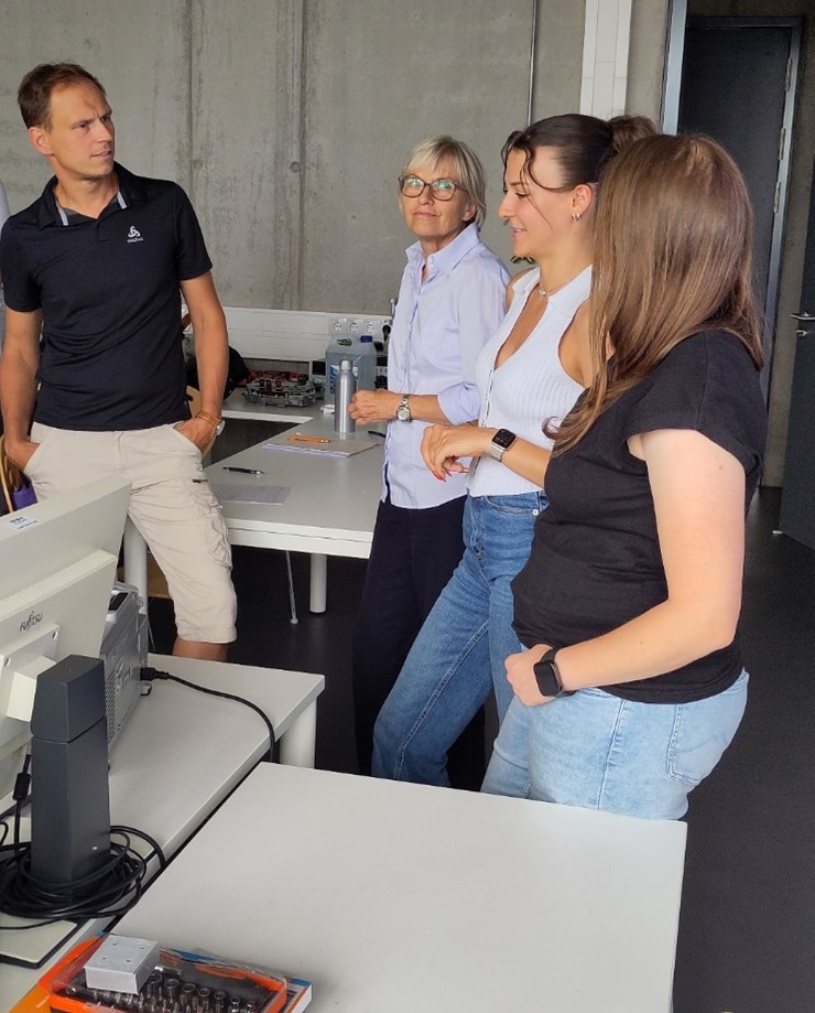 Vier Menschen stehen und reden in einem modernen, hellen Büro voller Schreibtische und elektronischer Geräte. Im Gespräch vertieft, diskutieren sie über technische Innovationen und nachhaltigen Wandel in ihrem gemeinschaftlichen Arbeitsbereich. at Hochschule Coburg