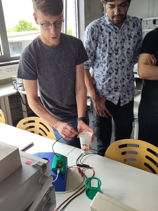 Zwei Personen stehen an einem Tisch mit elektronischen Geräten und erforschen technische Innovationen. Einer hält einen Pappbecher über ein Gerät, an dem Drähte angeschlossen sind, während der andere genau beobachtet. Im Hintergrund sind gelbe Stühle und große Fenster zu sehen. at Hochschule Coburg