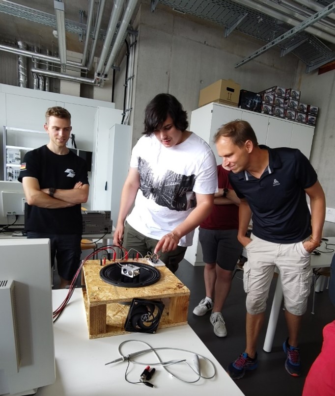Drei Personen stehen um ein hölzernes Gerät mit Drähten und Elektronik in einem Labor und diskutieren über technische Innovationen. Einer justiert das Gerät, während die anderen interessiert zusehen. Regale, Schränke und Kabel füllen den Hintergrund. at Hochschule Coburg
