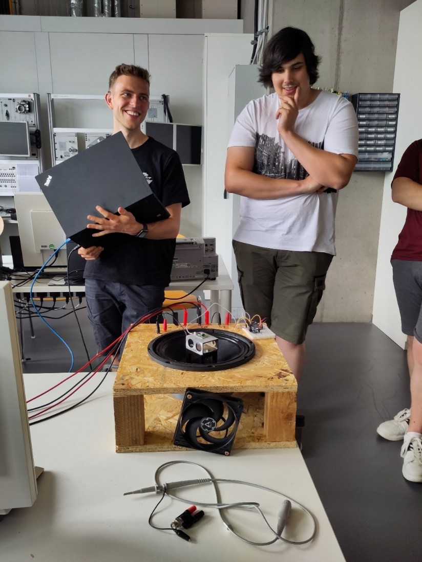 Zwei junge Männer stehen in einem Labor, einer hält einen Laptop in der Hand und lächelt. Vor ihnen steht eine Holzkiste mit einem großen Lautsprecher, Kabeln und einem Ventilator - ein technisches Projekt, das Technische Innovationen für nachhaltigen Wandel vorstellt. at Hochschule Coburg