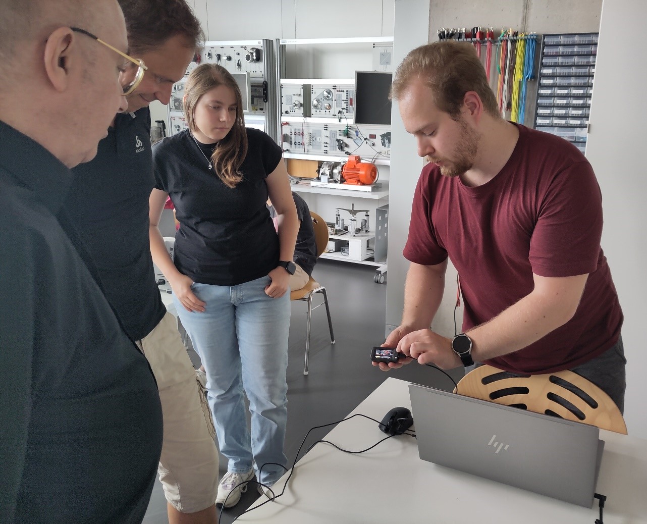 Ein Mann demonstriert drei Personen ein elektronisches Handgerät neben einem Laptop in einem modernen Klassenzimmer oder Labor, wobei technische Innovationen mit technischen Geräten im Hintergrund hervorgehoben werden. at Hochschule Coburg