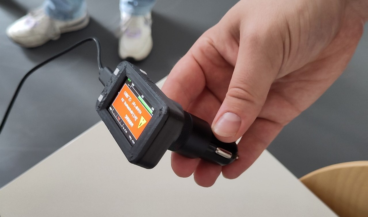 Eine Hand hält ein kleines elektronisches Gerät mit einem Bildschirm, der Text und ein Warnsymbol anzeigt - ein Beispiel für technische Innovationen; das Gerät ist an ein Kabel angeschlossen, im Hintergrund sind unscharfe Beine und Schuhe zu sehen. at Hochschule Coburg