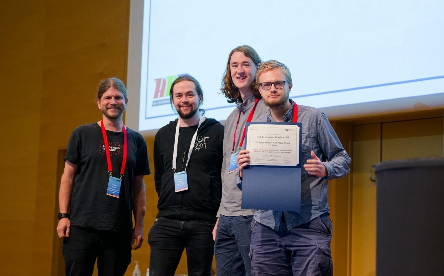 Vier junge Wissenschaftler stehen gemeinsam lächelnd vor einem großen Präsentationsbildschirm. Einer hält eine Urkunde in der Hand, mit der sie ihre Leistung im Visual-Computing feiern. Alle tragen Konferenz-Lanyards; die projizierten Namen sind über ihnen auf einer gelben Wand zu sehen. at Hochschule Coburg