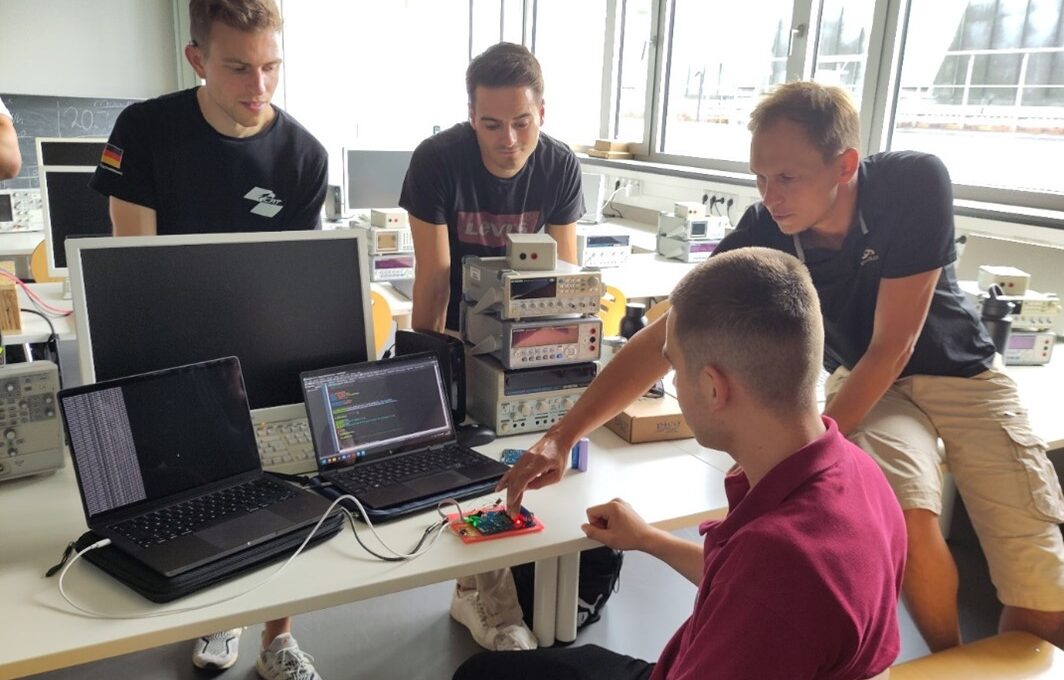Vier junge Männer arbeiten in einem hellen Klassenzimmer um einen Schreibtisch mit Laptops und elektronischen Geräten herum und diskutieren über technische Innovationen, während einer auf eine Platine zeigt, um nachhaltigen Wandel durch Teamarbeit und Kreativität voranzutreiben. at Hochschule Coburg