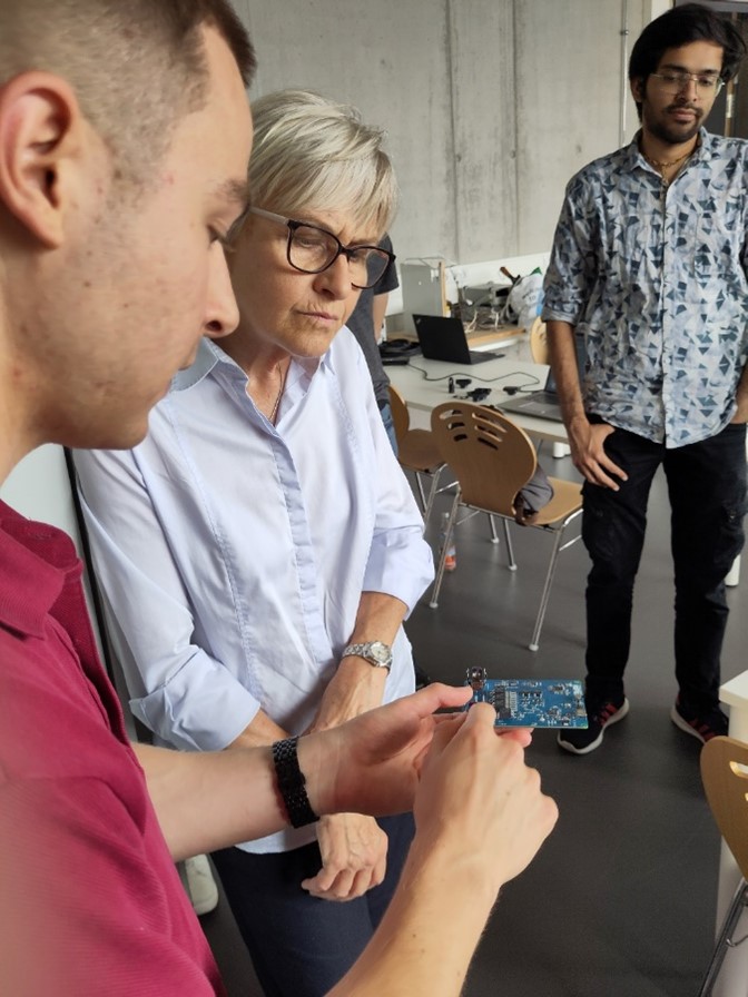 Ein Mann zeigt einer älteren Frau eine Platine, während ein anderer Mann in der Nähe in einem Klassenzimmer mit Holzstühlen und Laptops steht und auf technische Innovationen für nachhaltigen Wandel hinweist. at Hochschule Coburg