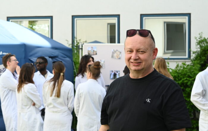Ein Mann mit schwarzem T-Shirt und Sonnenbrille steht lächelnd vor einer Gruppe von Menschen in weißen Laborkitteln, die sich im Freien in der Nähe eines Zeltes und einer Anzeigetafel versammelt haben, was auf eine Labor- oder Bioanalytik-Veranstaltung hindeutet. at Hochschule Coburg