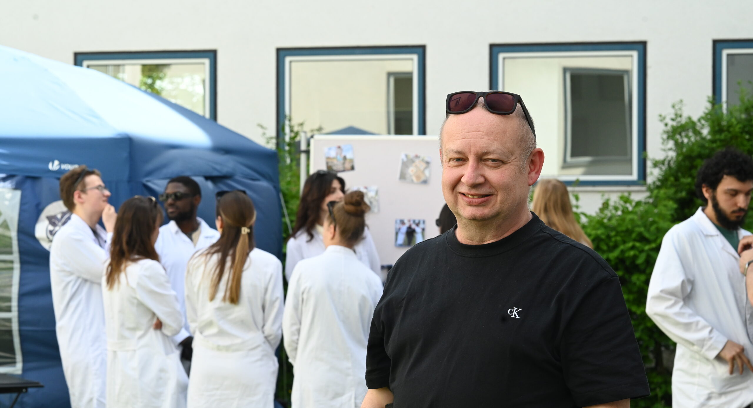 Ein Mann mit schwarzem T-Shirt und Sonnenbrille steht lächelnd vor einer Gruppe von Menschen in weißen Laborkitteln, die sich im Freien in der Nähe eines Zeltes und einer Anzeigetafel versammelt haben, was auf eine Labor- oder Bioanalytik-Veranstaltung hindeutet. at Hochschule Coburg