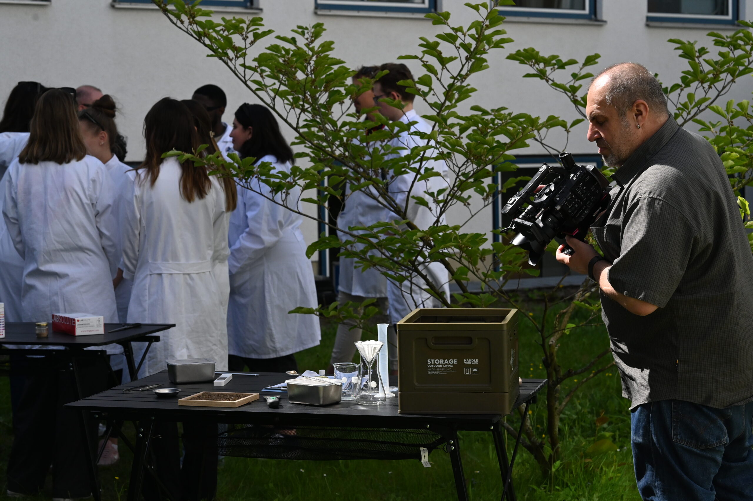 Ein Mann filmt eine Gruppe von Personen in weißen Laborkitteln, die im Freien versammelt sind und möglicherweise Bioanalytik-Experimente durchführen. Im Vordergrund ist ein Tisch mit Laborgeräten und einer grünen Aufbewahrungsbox zu sehen. Auch Bäume und ein Gebäude sind zu sehen. at Hochschule Coburg