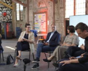 Six people sit in a row on a stage with exposed brick walls and a mural. One woman on the left moderates the engaging panel discussion at Impulse Coburg, part of the upcoming Nachhaltigkeitstage 2025 event series. at Hochschule Coburg