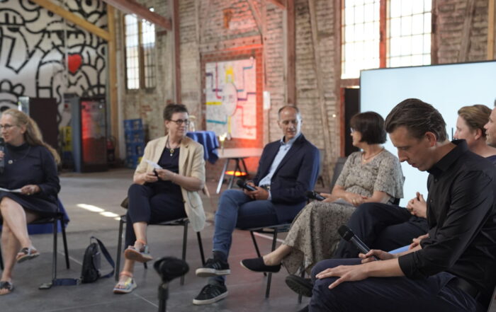 Six people sit in a row on a stage with exposed brick walls and a mural. One woman on the left moderates the engaging panel discussion at Impulse Coburg, part of the upcoming Nachhaltigkeitstage 2025 event series. at Hochschule Coburg