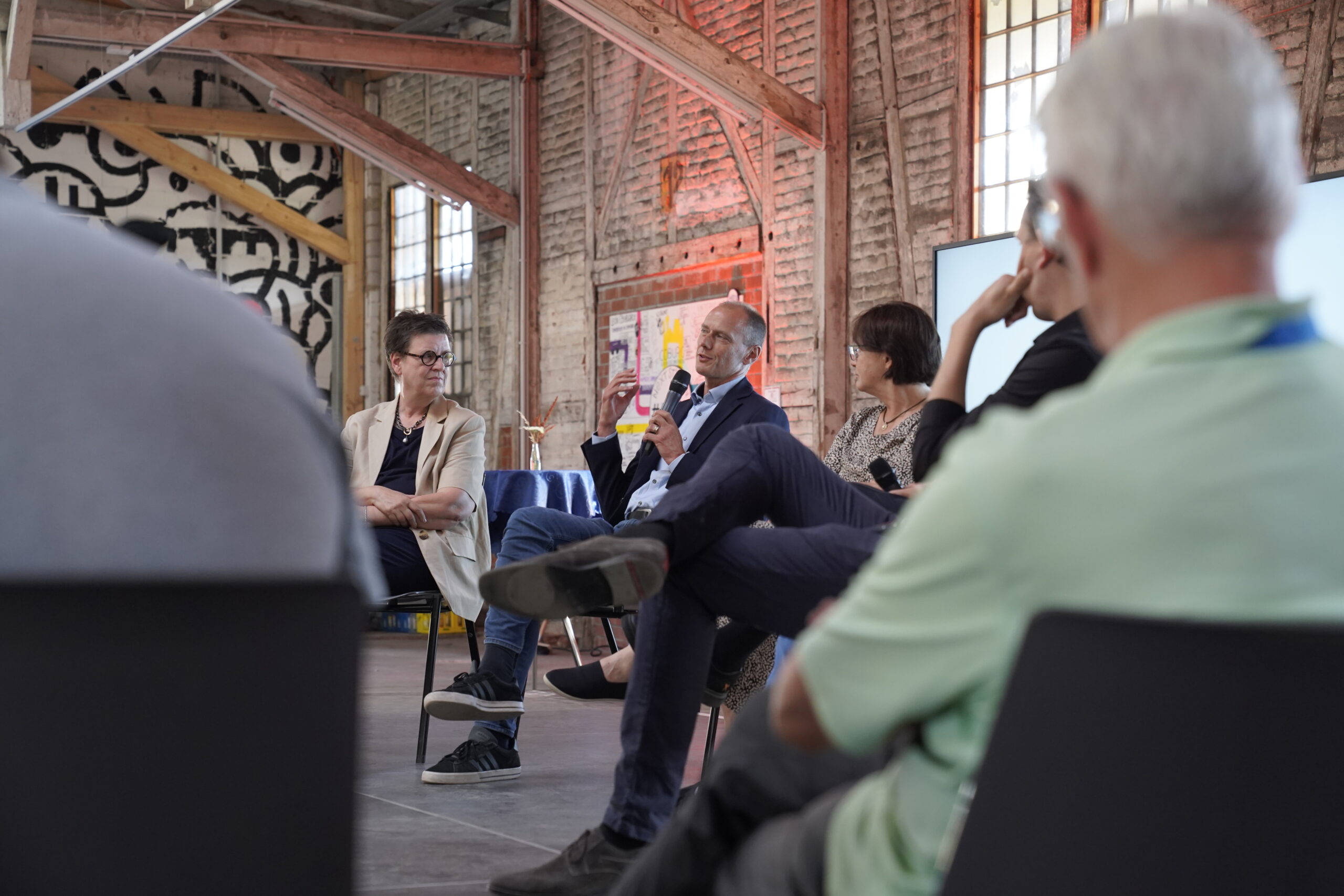Eine Gruppe von Menschen sitzt im Kreis und diskutiert in einem Raum mit freiliegenden Backsteinwänden und großen Fenstern. Ein Mann im Anzug spricht, während die anderen aufmerksam zuhören. at Hochschule Coburg