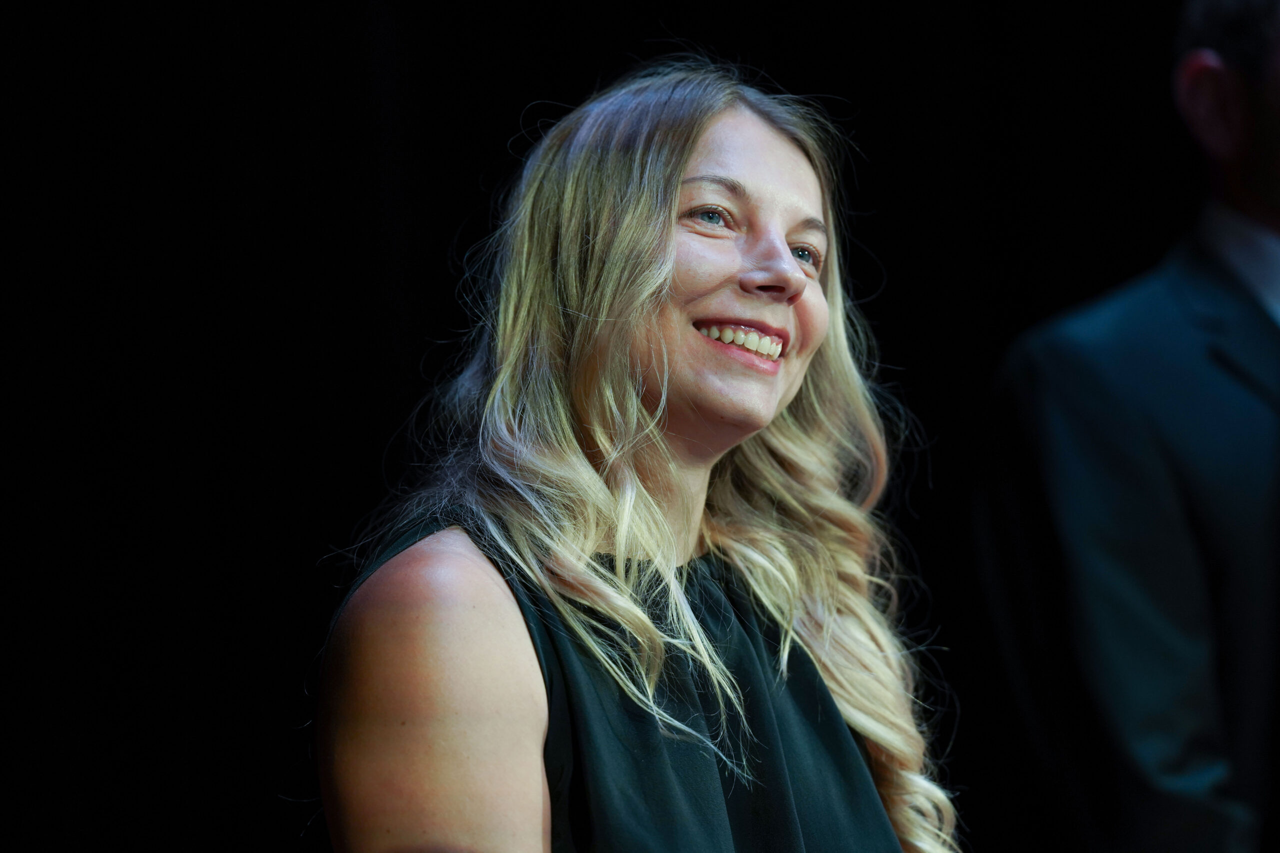Tetyana Hryhorenko, mit langen, gewellten blonden Haaren und einem ärmellosen schwarzen Oberteil, lächelt, während sie vor einem dunklen Hintergrund leicht nach oben blickt - und strahlt das Selbstbewusstsein einer DAAD-Preisträgerin 2025 aus. at Hochschule Coburg