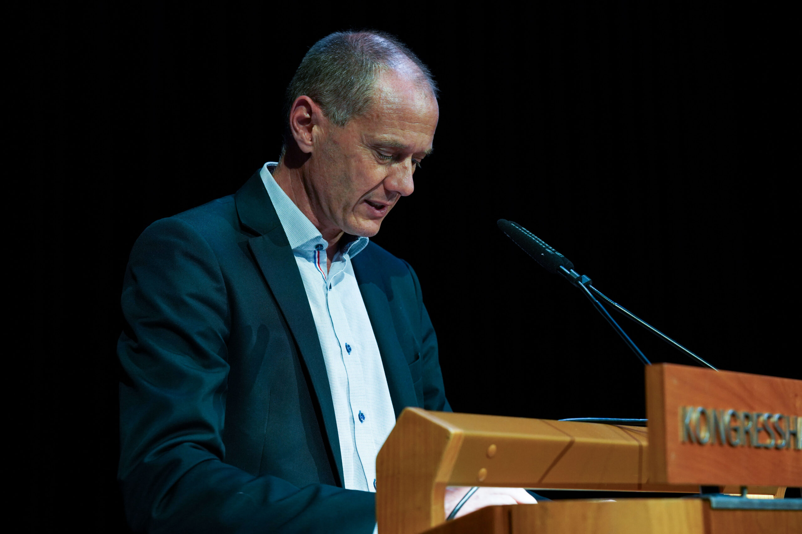 Ein Mann im Anzug steht an einem hölzernen Podium, spricht oder liest, vor ihm ein Mikrofon, dahinter ein dunkler Hintergrund. at Hochschule Coburg