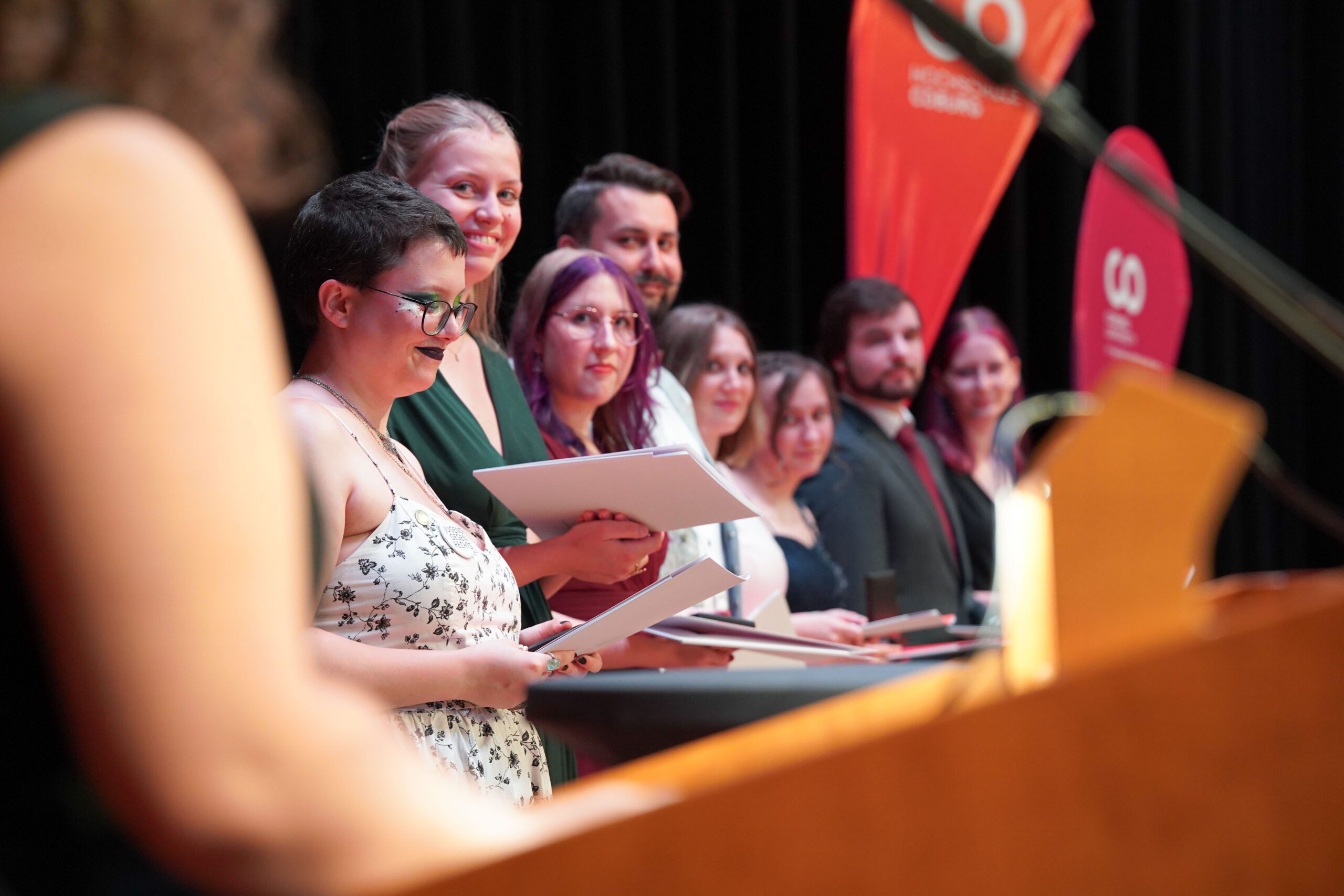Eine Gruppe von Menschen steht in einer Reihe auf der Bühne, hält Urkunden und lächelt. Eine Person auf einem Podium ist im Vordergrund unscharf zu erkennen. Im Hintergrund sind rote Fahnen zu sehen. at Hochschule Coburg