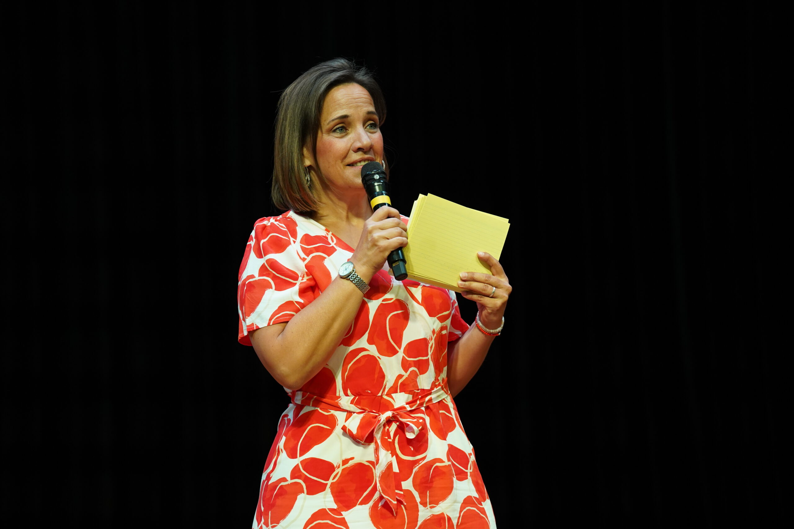 Bei der feierlichen Absolventenverabschiedung 2025 spricht eine Frau in einem rot-weiß gemusterten Kleid in ein Mikrofon, während sie einen Notizblock in der Hand hält und vor einem schwarzen Hintergrund steht, wodurch ein unvergesslicher Moment entsteht. at Hochschule Coburg