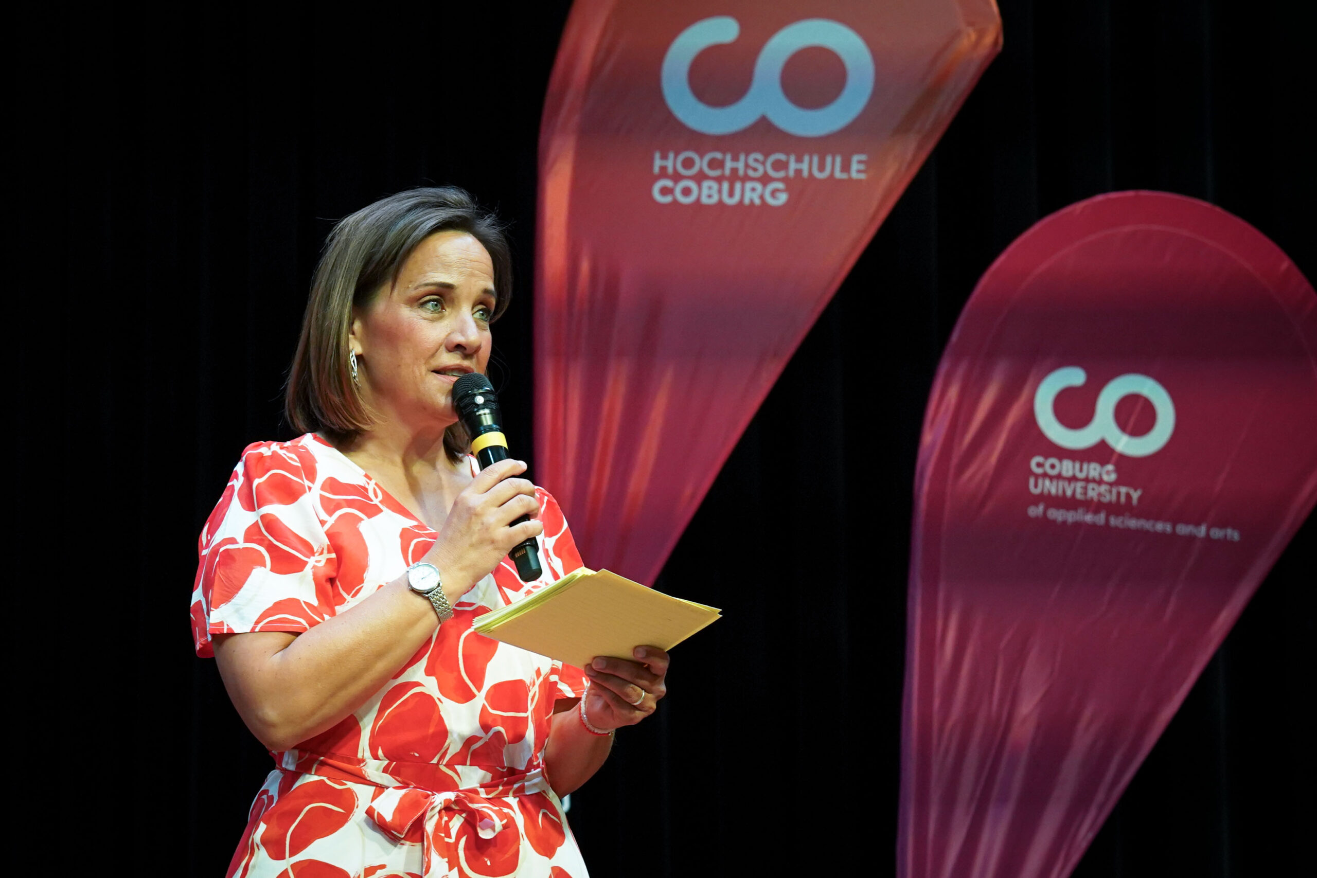 Eine Frau in einem rot-weißen Kleid spricht in ein Mikrofon und hält Papiere in der Hand. Hinter ihr sind zwei rote Banner mit den Logos und Namen der Hochschule Coburg und der Fachhochschule Coburg zu sehen. at Hochschule Coburg
