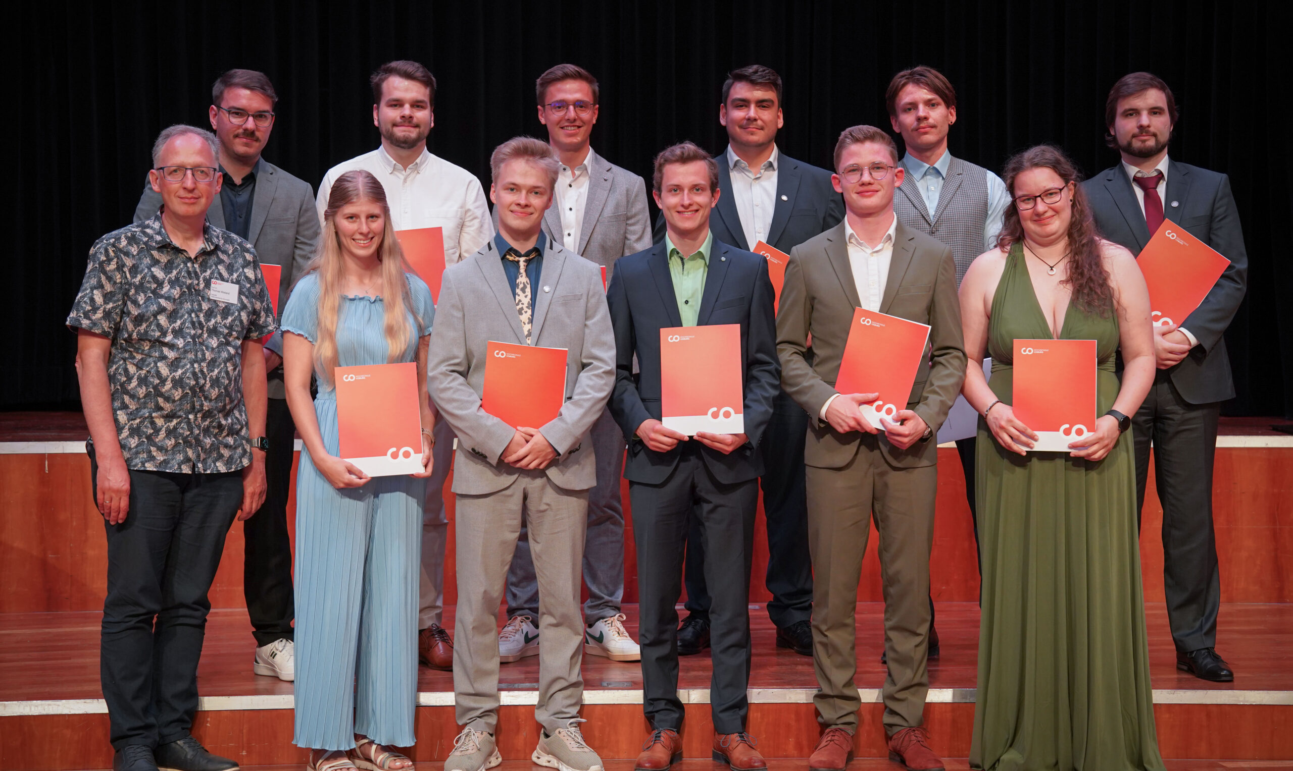 Eine Gruppe von 14 Personen, zumeist junge Erwachsene, die förmlich gekleidet sind, stehen auf einer Bühne, halten rote Mappen und lächeln in die Kamera. Ein Mann auf der linken Seite trägt ein gemustertes Hemd; der Hintergrund ist ein dunkler Vorhang. at Hochschule Coburg