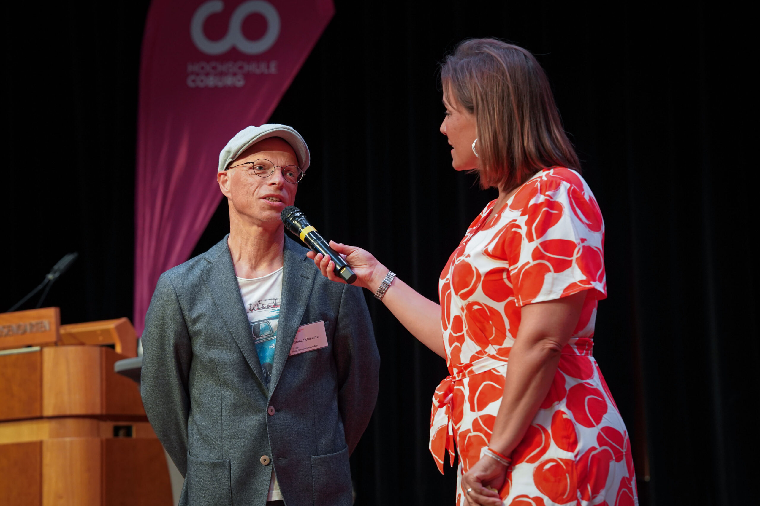 Eine Frau in einem rot-weißen Kleid interviewt einen Mann in einem grauen Blazer und einer Mütze auf der Bühne. Die Frau hält ein Mikrofon in der Hand. Im Hintergrund sind ein rosa Banner und ein Holzpodium zu sehen. at Hochschule Coburg