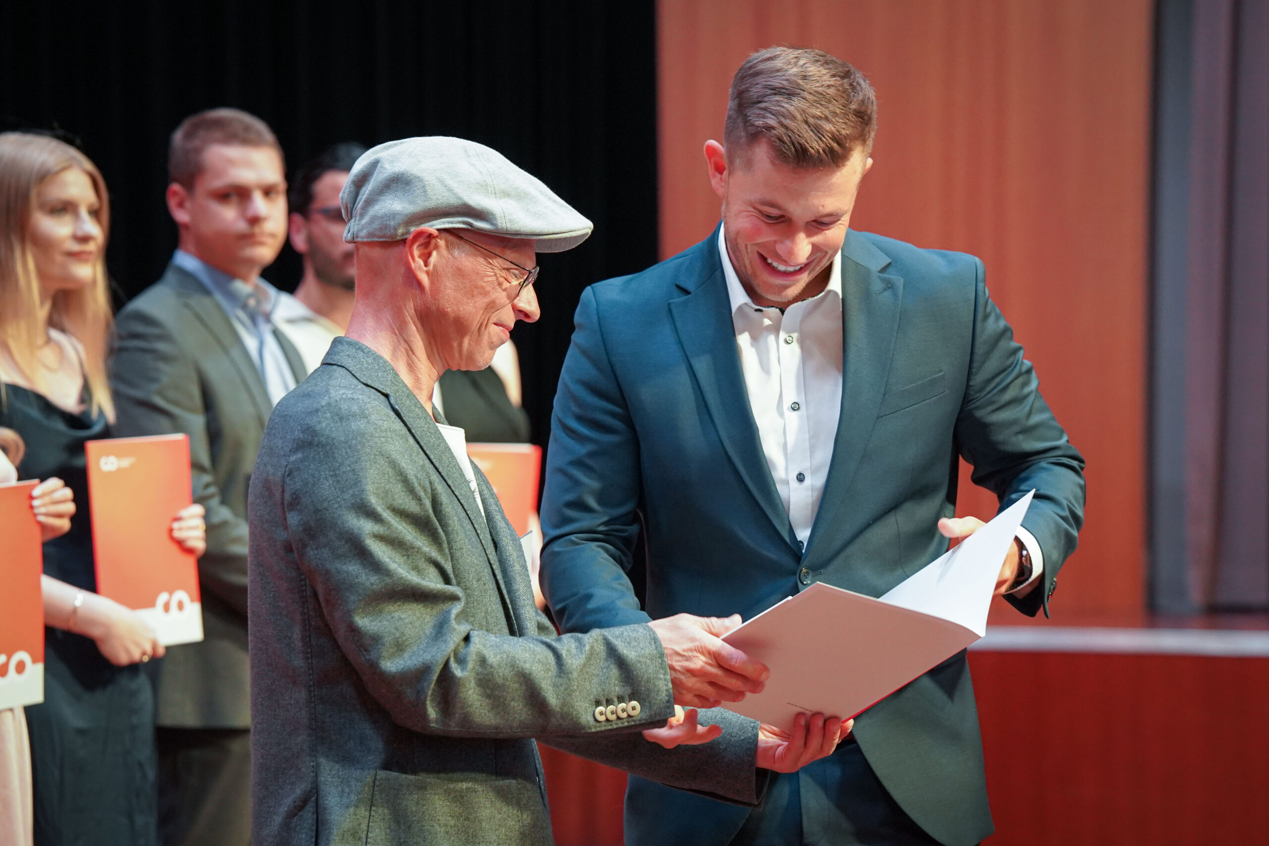Ein lächelnder Mann im Anzug erhält während einer Preisverleihung eine Urkunde von einem älteren Mann mit Mütze und Brille, während andere Personen im Hintergrund Dokumente halten. at Hochschule Coburg
