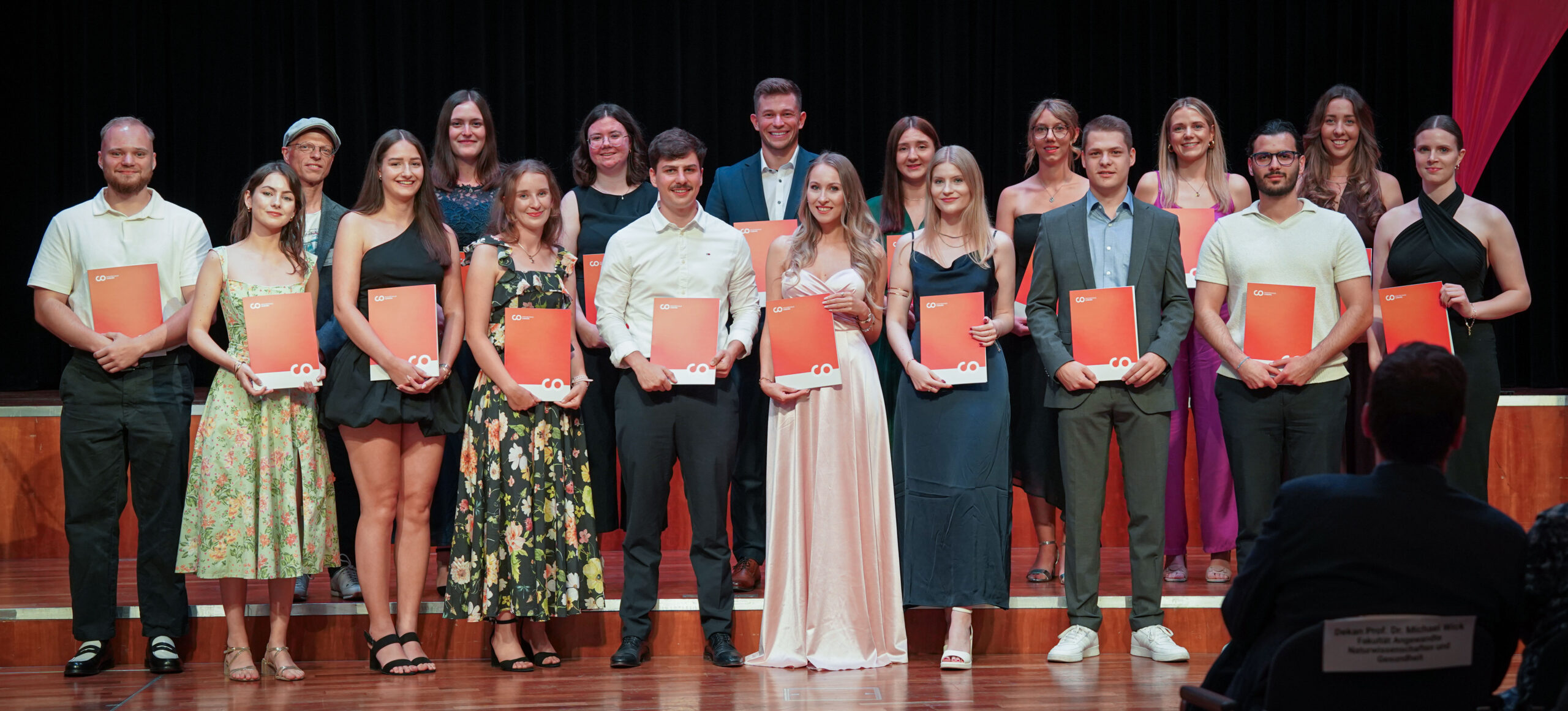 Eine Gruppe junger Erwachsener in formeller Kleidung steht auf einer Bühne, lächelt und hält orangefarbene Urkunden in der Hand. Sie posieren für ein Foto bei einer Abschluss- oder Preisverleihungsfeier. Im Hintergrund sind schwarze Vorhänge und das Publikum zu sehen. at Hochschule Coburg