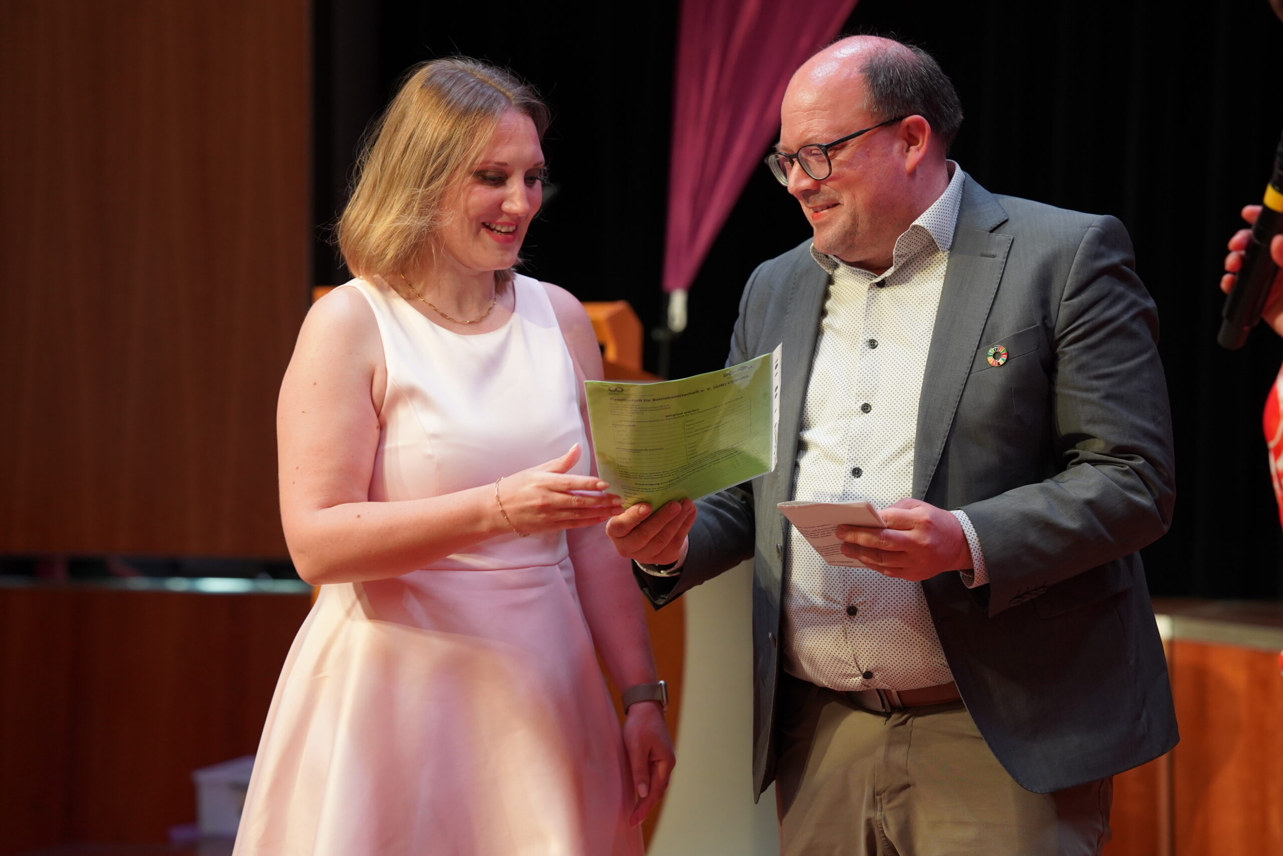 Bei der feierlichen Absolventenverabschiedung 2025 tauschen eine Frau in einem hellrosa Kleid und ein Mann in einem grauen Anzug auf der Bühne eine grüne Urkunde aus, beide lächeln, als sie diesen unvergesslichen Moment vor verschwommenen lila Vorhängen teilen. at Hochschule Coburg