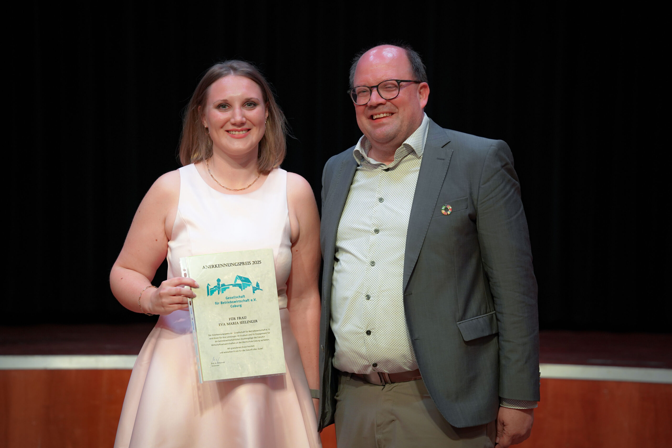 Eine Frau in einem hellrosa Kleid hält eine Urkunde in der Hand und steht neben einem lächelnden Mann in einer Anzugsjacke. Beide posieren auf einer Bühne mit einem dunklen Vorhang im Hintergrund. at Hochschule Coburg