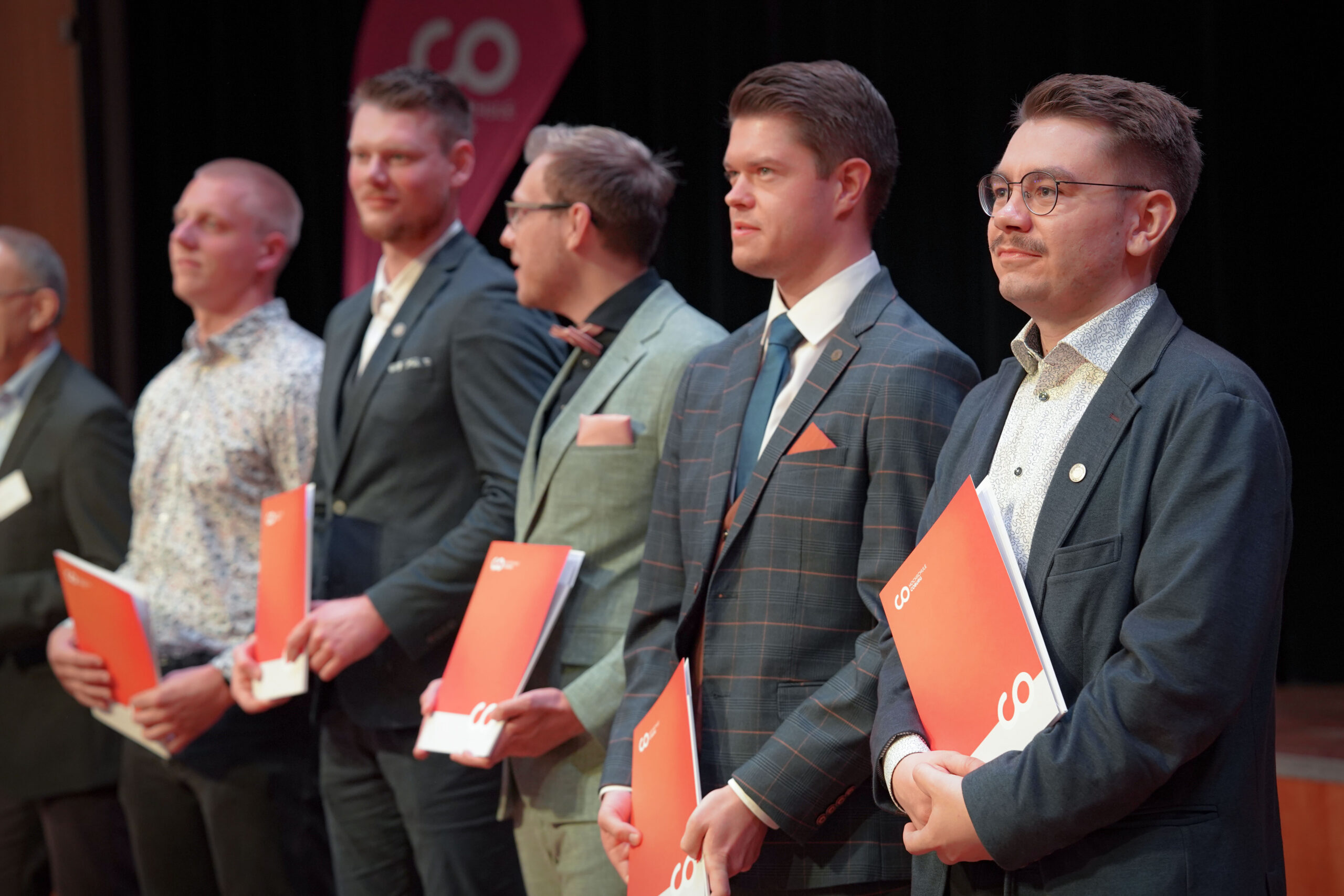 Eine Gruppe von Männern in formeller Kleidung steht in einer Reihe auf der Bühne, jeder hält eine orangefarbene Mappe. Sie scheinen an einer Veranstaltung oder Zeremonie teilzunehmen, mit einem dunklen Hintergrund und einem teilweise sichtbaren Logo hinter ihnen. at Hochschule Coburg