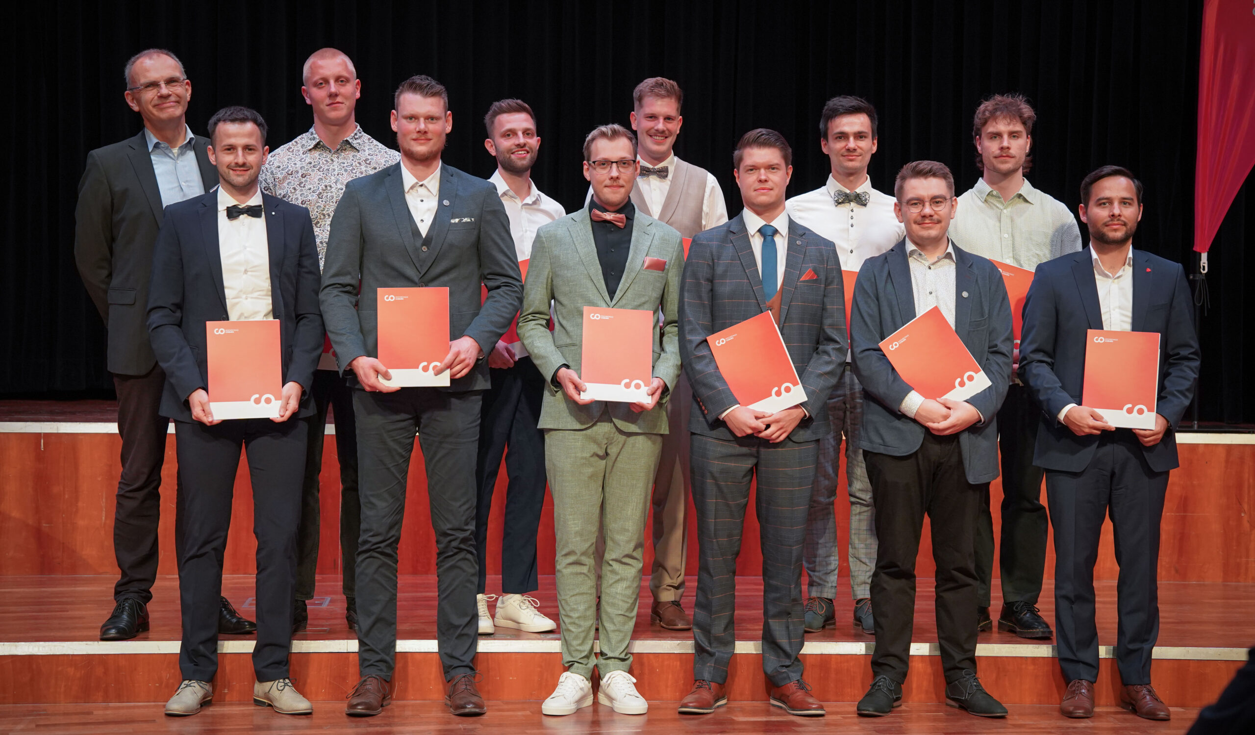 Dreizehn Männer in formeller Kleidung stehen auf einer Bühne und posieren für ein Gruppenfoto. In der ersten Reihe stehen rote Urkunden oder Mappen. Im Hintergrund sind ein dunkler Vorhang und rote Dekorationen zu sehen. at Hochschule Coburg
