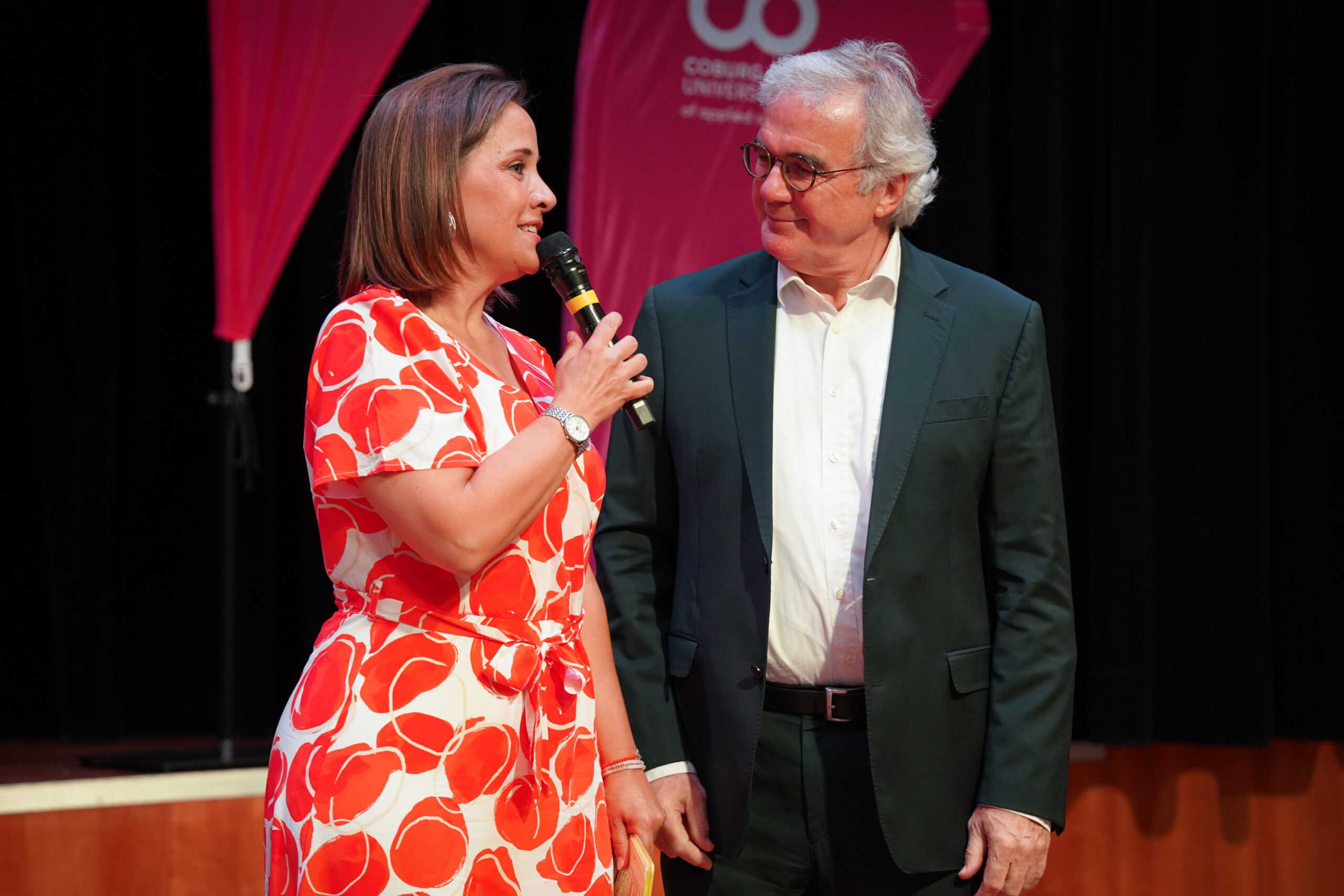 Eine Frau in einem rot-weißen Kleid spricht in ein Mikrofon, während sie neben einem Mann in einem dunklen Anzug steht; beide befinden sich auf einer Bühne mit einem rosa Banner und einem dunklen Hintergrund. at Hochschule Coburg