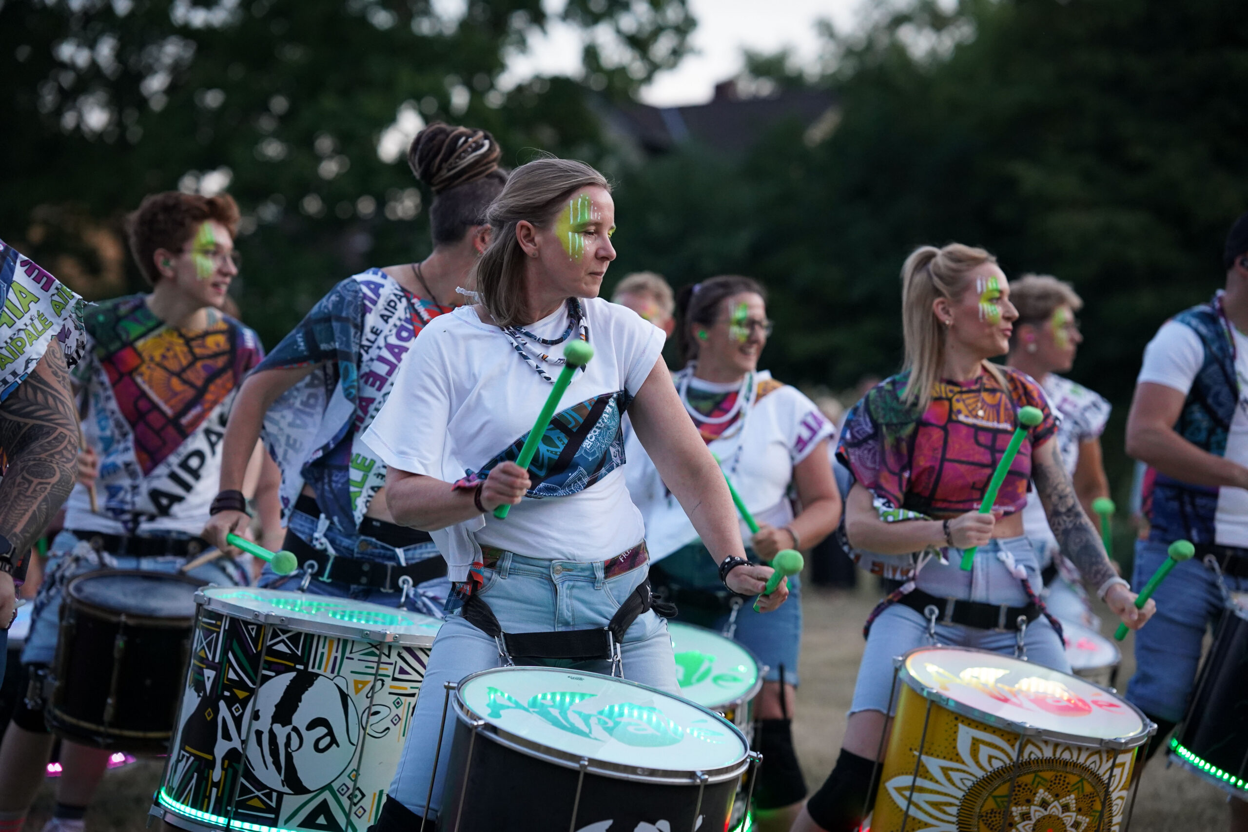 Eine Gruppe von Menschen spielt im Freien auf Trommeln und trägt dabei bunte, gemusterte Kleidung und Gesichtsbemalung. Die Trommeln haben lebendige Muster und LED-Lichter. Im Hintergrund sind Bäume und unscharfe Gebäude zu sehen. at Hochschule Coburg