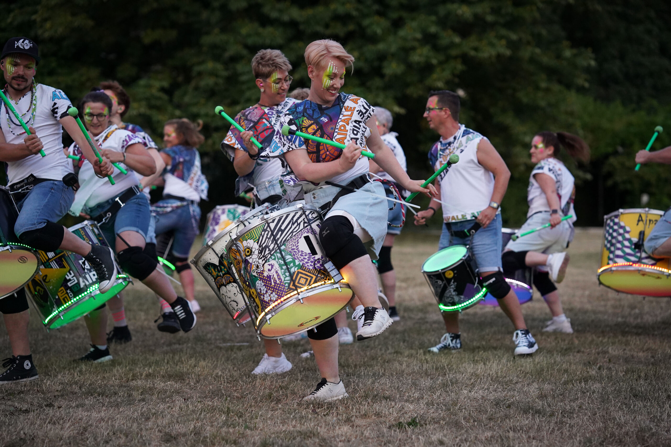 Eine Gruppe von Menschen spielt im Freien auf dekorierten Trommeln, sie tragen bunte Schminke und legere Kleidung. Sie tanzen und trommeln aktiv auf einer Wiese mit Bäumen im Hintergrund und strahlen Energie und Freude aus. at Hochschule Coburg