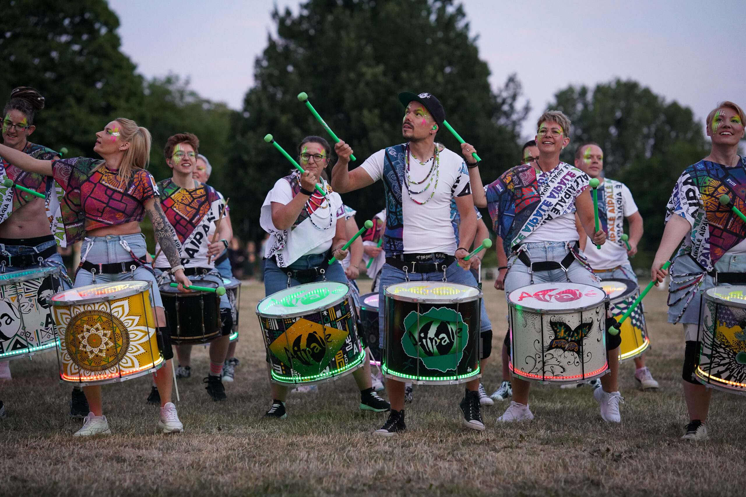 Eine Gruppe von Menschen in farbenfroher Kleidung und mit Gesichtsbemalung spielt auf großen geschmückten Trommeln im Freien auf einer Wiese. Im Hintergrund sind Bäume zu sehen und die Gruppe wirkt energiegeladen und fröhlich. at Hochschule Coburg