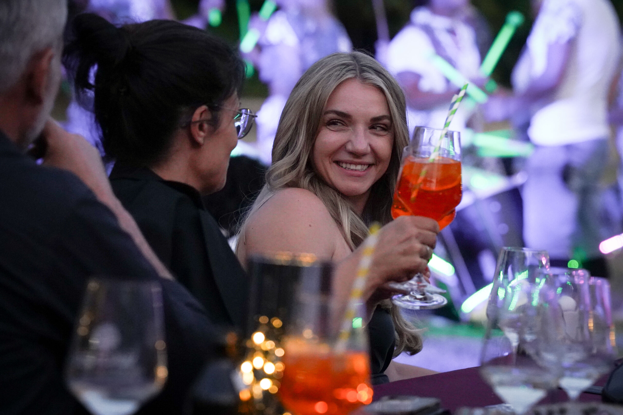 Eine Frau mit langen blonden Haaren lächelt und hebt ein Cocktailglas, während sie mit anderen an einem Tisch im Freien sitzt, umgeben von Getränken und weichen, bunten Lichtern im Hintergrund. at Hochschule Coburg