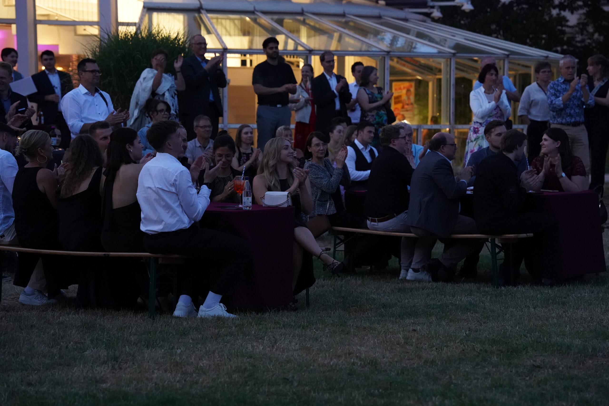 Eine Gruppe von Menschen sitzt und steht in der Abenddämmerung um Tische im Freien und beobachtet eine feierliche Veranstaltung. Die Tische sind mit weinroten Tüchern bedeckt, und die Gäste in halb-formeller Kleidung feiern die unvergessliche Absolventenverabschiedung 2025, während im Hintergrund eine Glaskonstruktion glänzt. at Hochschule Coburg