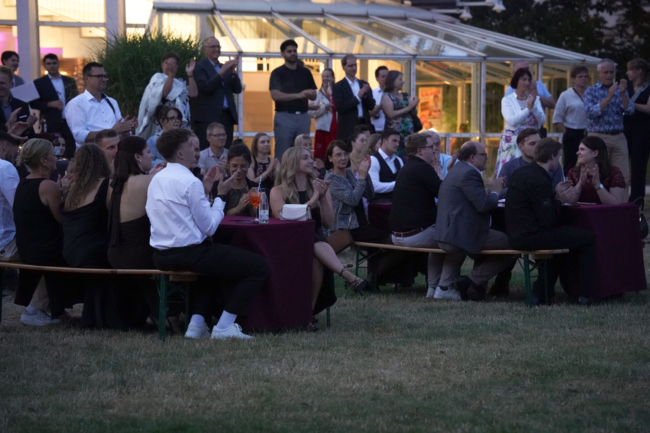 Eine Gruppe von Menschen sitzt an Tischen und Bänken im Freien, klatscht und beobachtet eine Veranstaltung, während andere in der Abenddämmerung im Hintergrund neben einem Glasgebäude stehen. at Hochschule Coburg