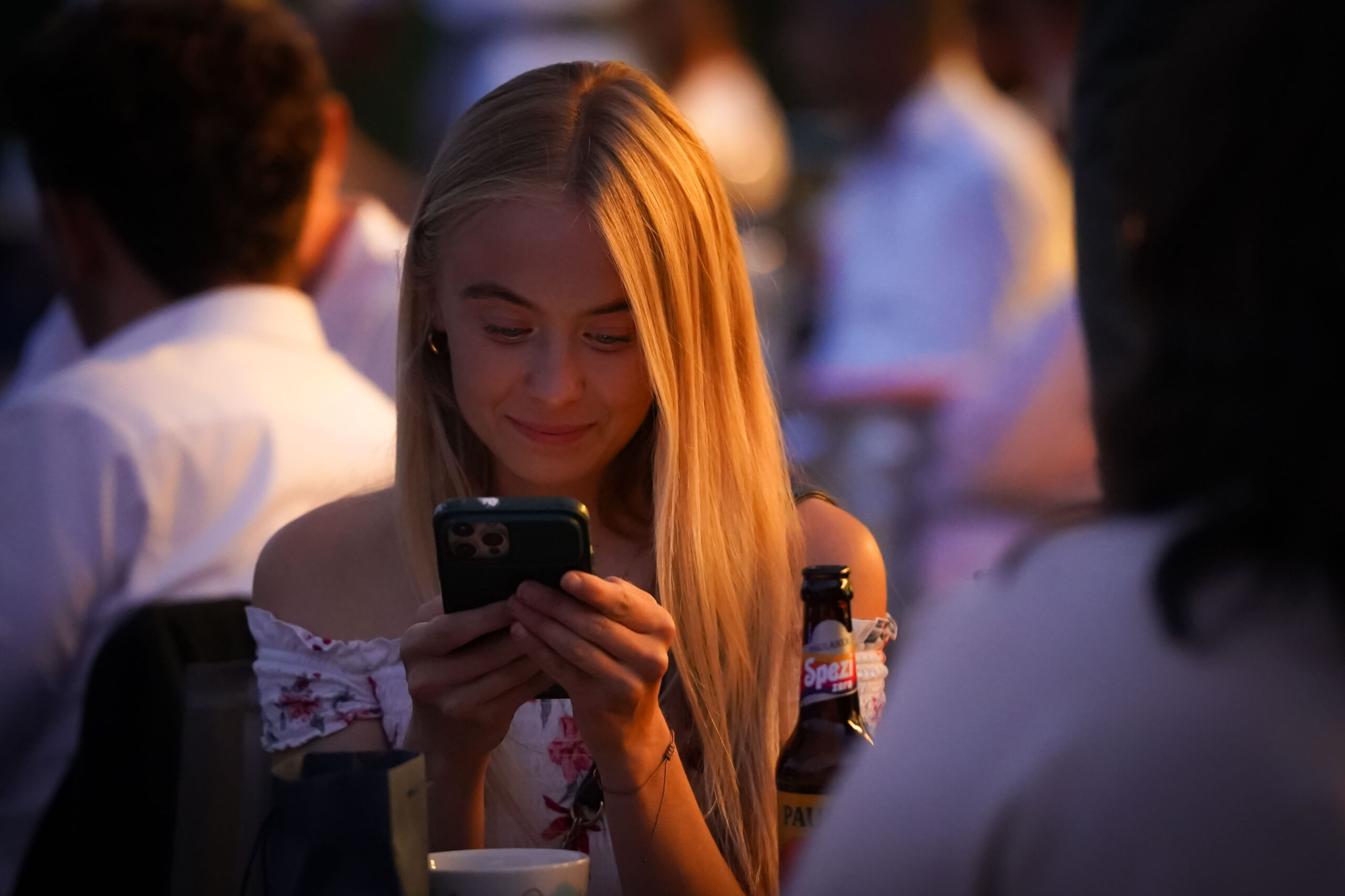 Eine junge Frau mit langen blonden Haaren lächelt, während sie bei einem Treffen im Freien auf ihr Telefon schaut, mit warmer Beleuchtung und unscharfen Menschen im Hintergrund. Eine Flasche und eine Tasse stehen auf dem Tisch vor ihr. at Hochschule Coburg